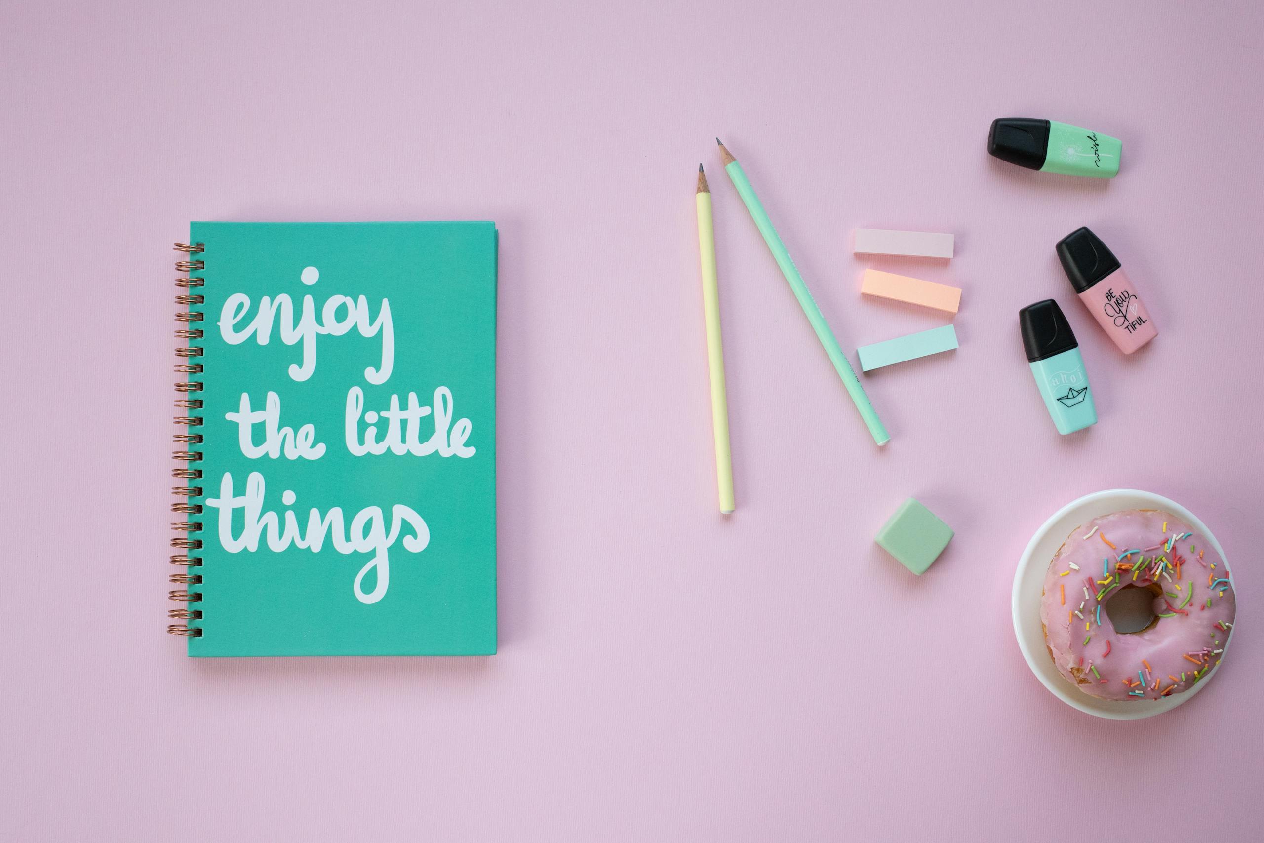 A vibrant flat lay of desk items with a positive message notebook and colorful donut on a pink background.
