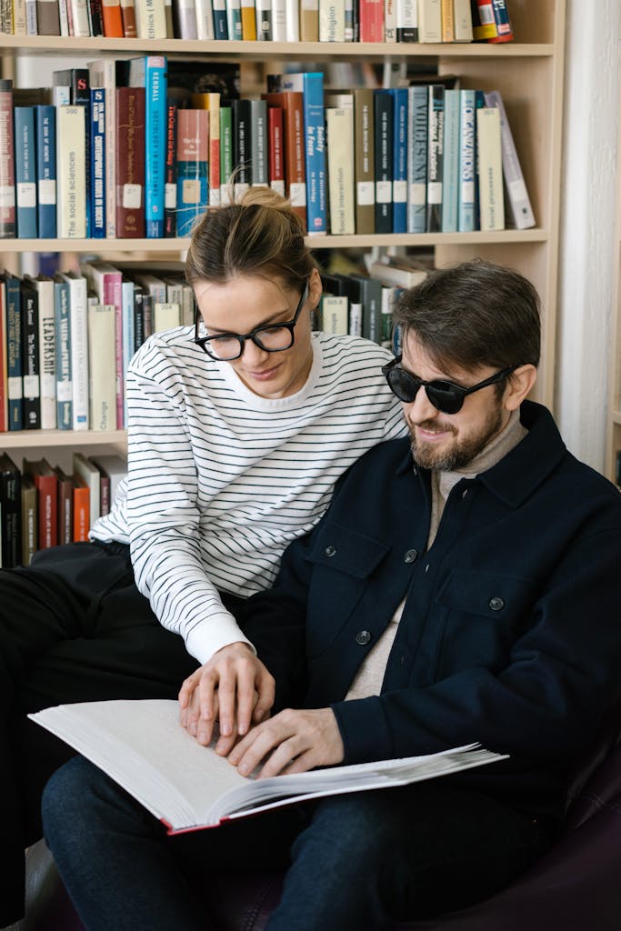 A woman helps a visually impaired man read a Braille book in a library setting.